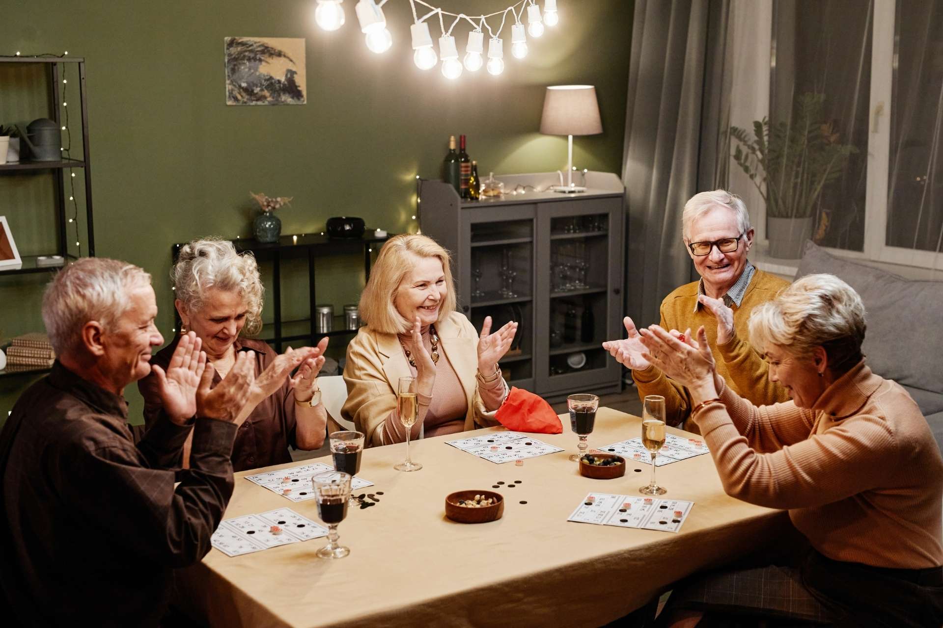 A group of older adults gathered around a table, laughing and clapping while playing a game together in a cozy, warmly lit living room with drinks and snacks.